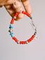 Scarlet Maverick bracelet held between fingers, showing red colored-glaze beads, turquoise accents, a fishhook clasp and a round charm.