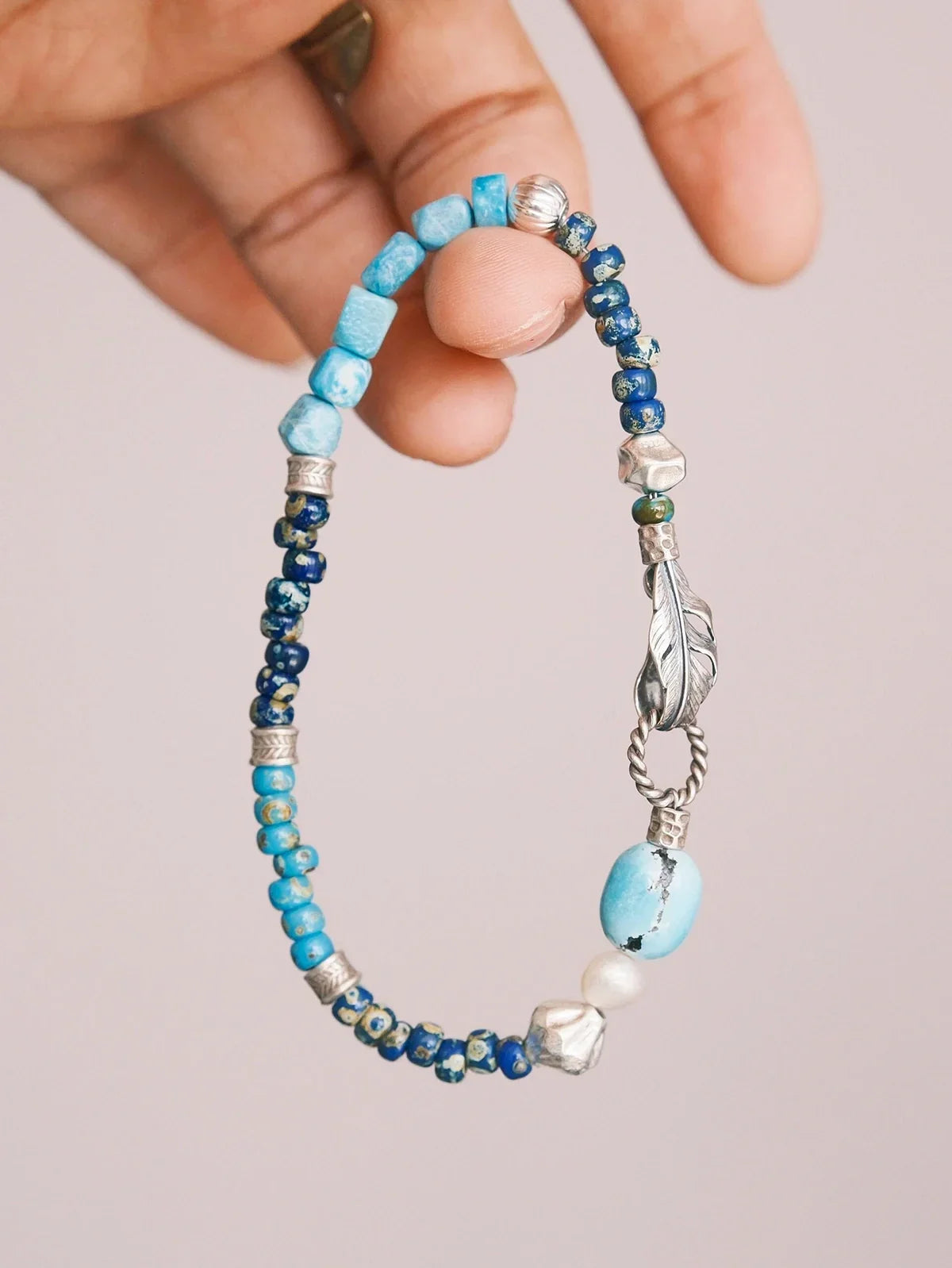 Blue Plume bracelet held in a hand, showing blue beads, a pearl accent, textured silver spacers and a feather clasp.