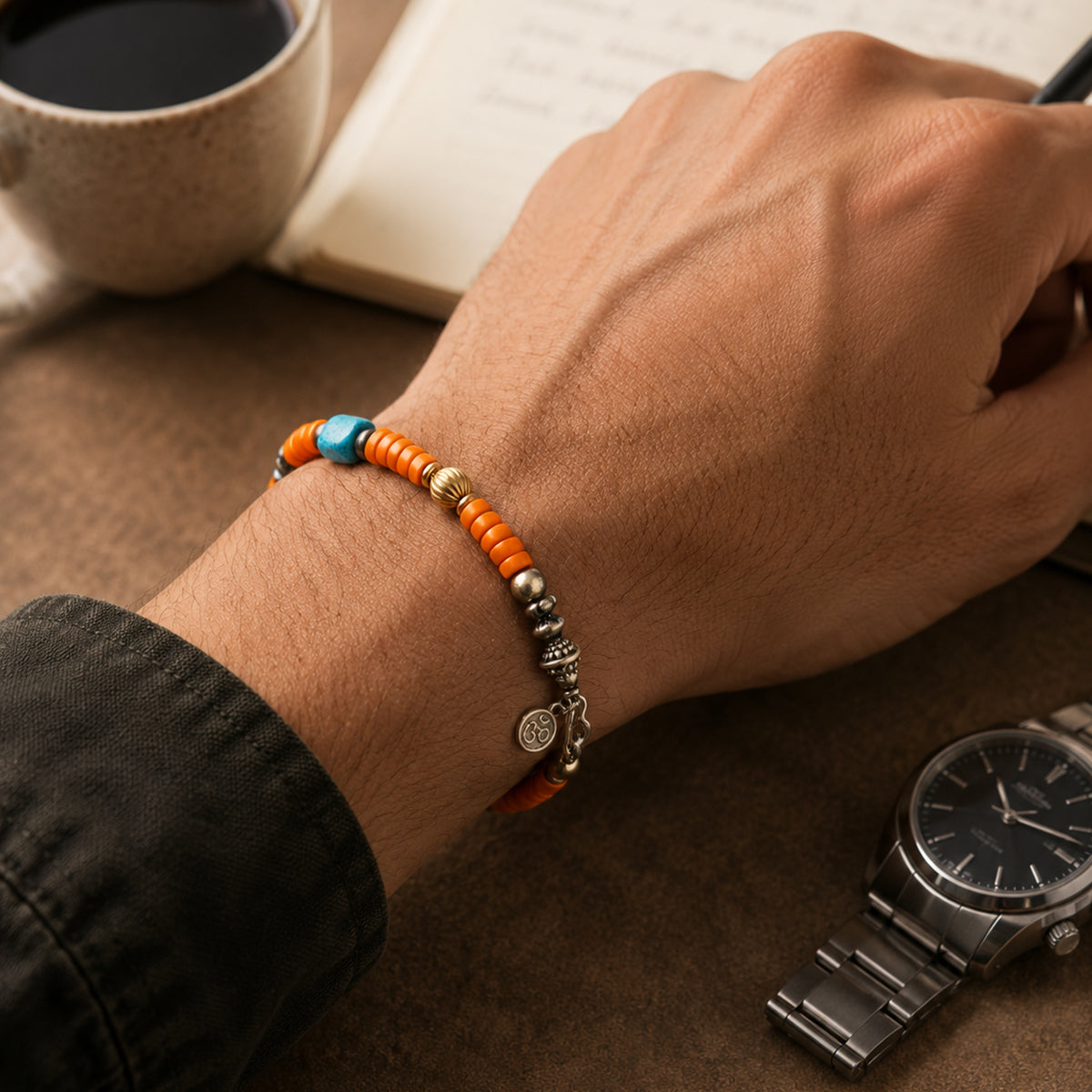 Close-up of a wrist wearing an orange and turquoise-blue beaded bracelet beside a notebook, coffee cup and watch.