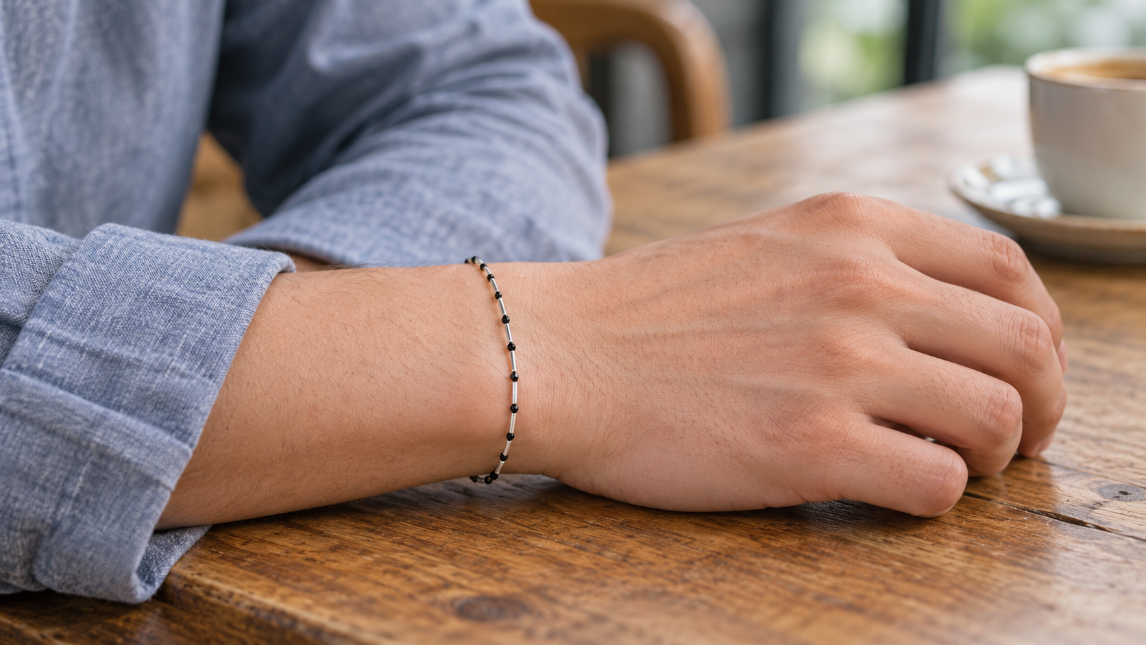 A slim black and silver-colored bracelet worn with a blue shirt at a wooden table.