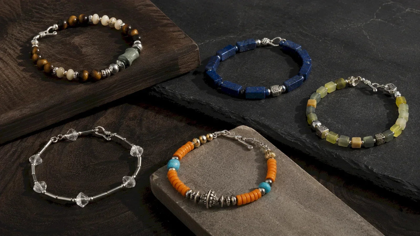 Five ASIA'NOBLE men's bracelets in tiger eye, lapis lazuli, multicolor jade, clear quartz and turquoise displayed on dark wood and slate.