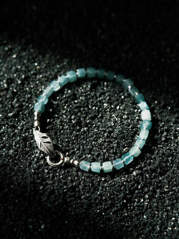 Blue jade bracelet for men displayed on a dark textured surface, sterling silver feather jewelry with calm modern contrast