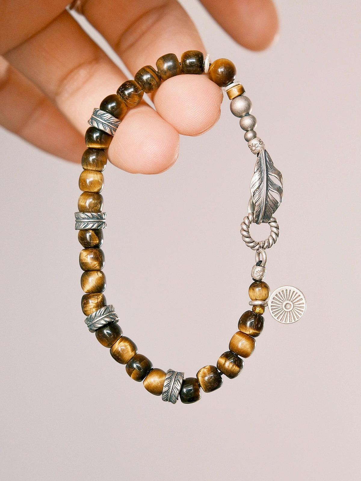Hand holding a tiger eye men’s bracelet with sterling silver feather charm, close-up product angle, refined natural stone jewelry. Main image