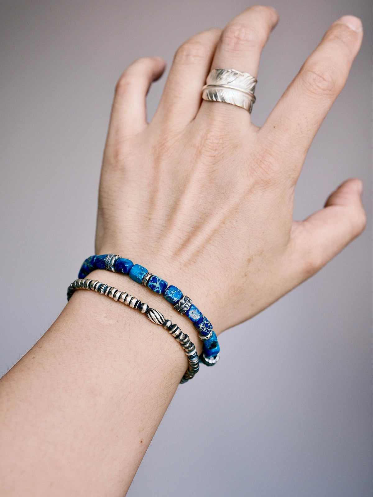 Men's silver feather bracelet stacked with a blue lapis-style bracelet on wrist, casual layered jewelry styling