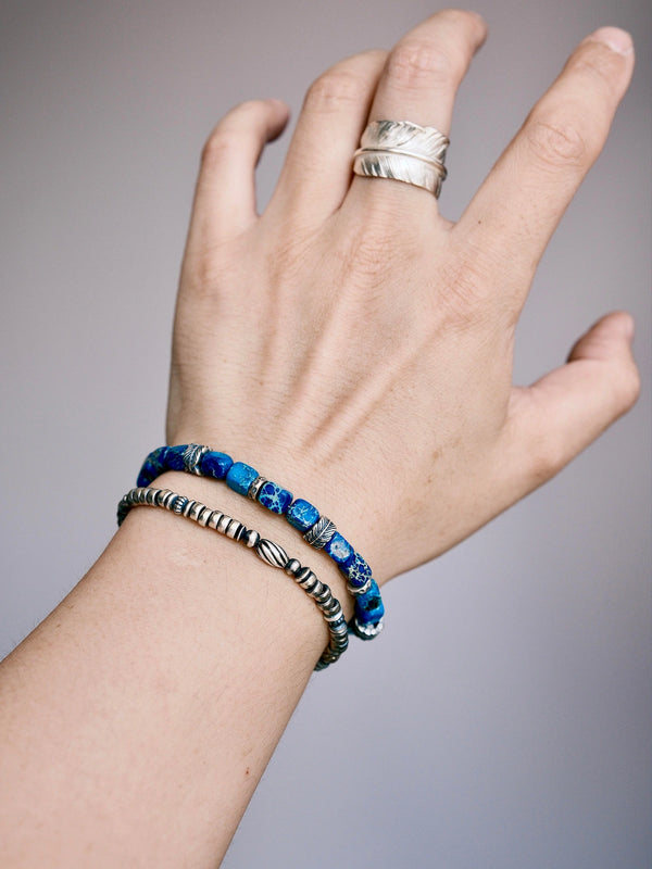 Men's silver feather bracelet stacked with a blue lapis-style bracelet on wrist, casual layered jewelry styling