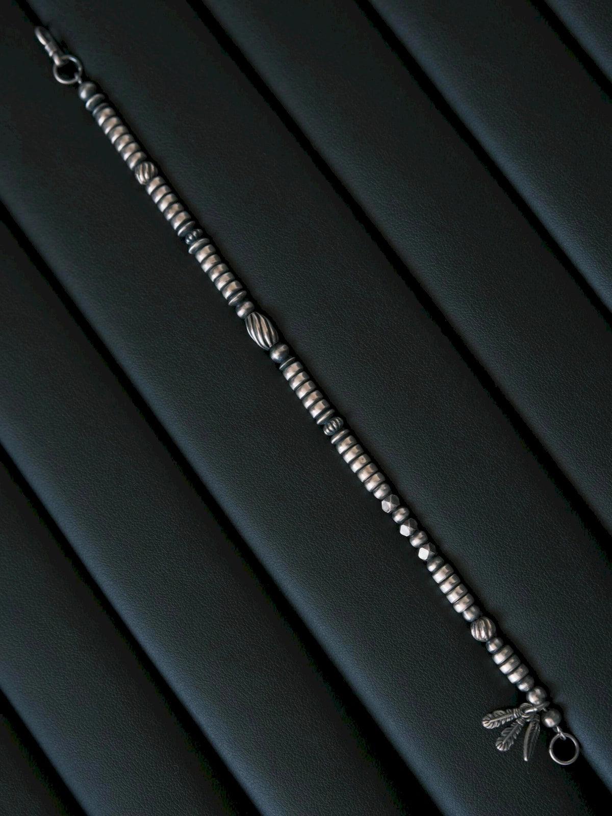 Full-length Silver Nomad sterling silver bracelet laid on black leather, oxidized silver beads and feather charm detail