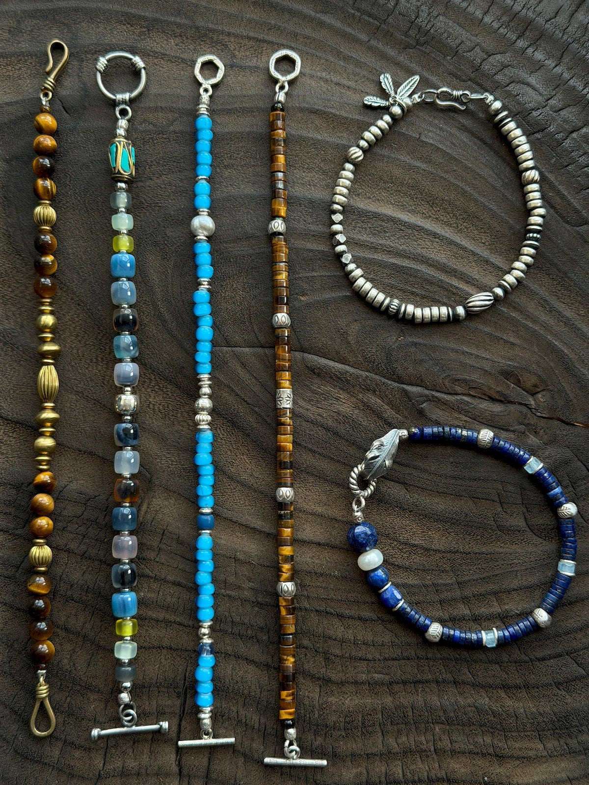 Silver Nomad men's silver feather bracelet displayed with mixed gemstone bracelets on dark carved wood, Identity Series flat lay