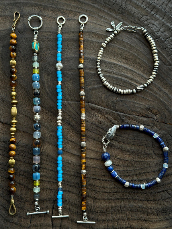 Silver Nomad men's silver feather bracelet displayed with mixed gemstone bracelets on dark carved wood, Identity Series flat lay