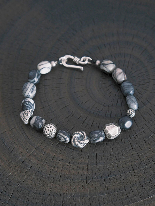 Flat lay of men’s black and white webbed jasper-style stone bracelet with heavy sterling silver accents on dark wood, urban stoic style.