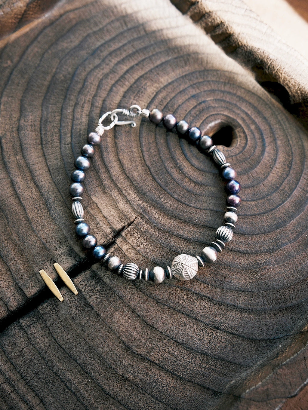 Men’s natural pearl and hammered silver bracelet laid flat on dark wood, cold ocean tones, refined masculine jewelry.