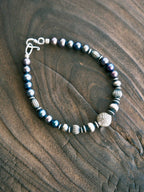 Men’s natural pearl and hammered silver bracelet laid flat on dark wood, cold ocean tones, refined masculine jewelry.