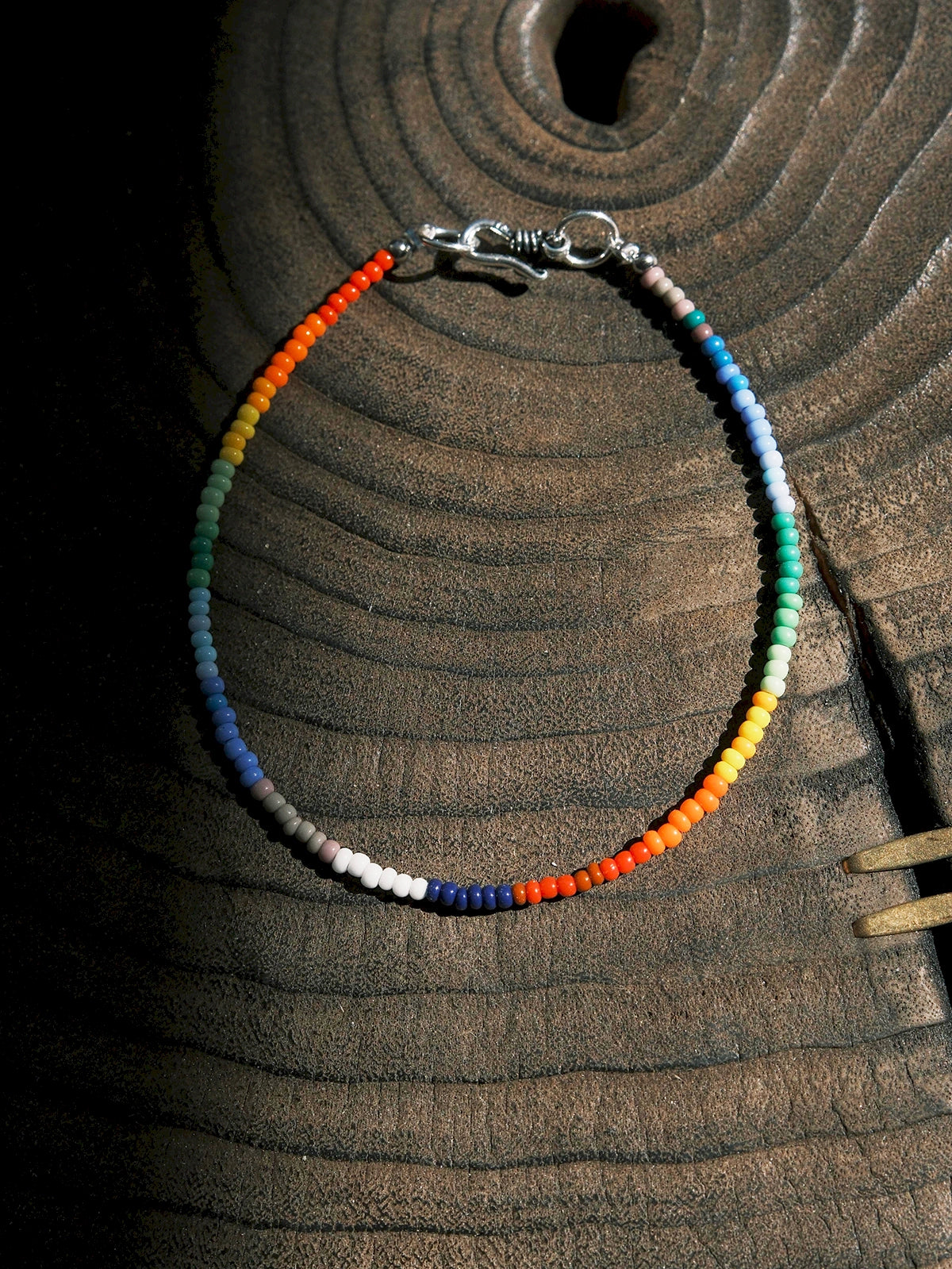 Ultra-thin rainbow colored-glaze men’s bracelet with sterling silver clasp, flat lay on dark textured background, minimal identity style