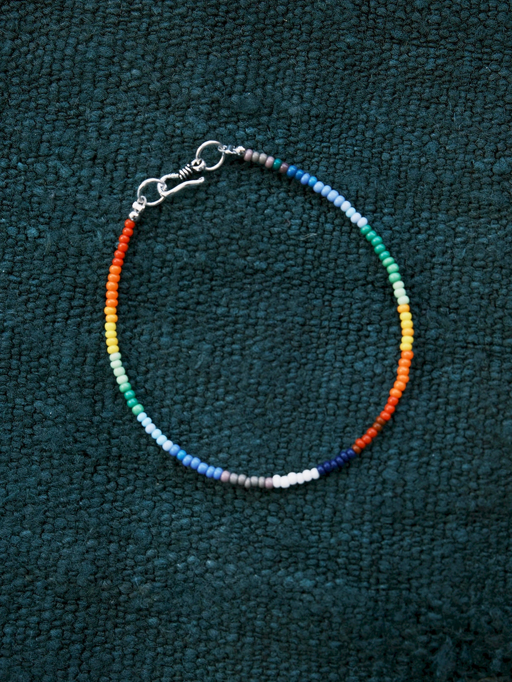 Ultra-thin rainbow colored-glaze men’s bracelet with sterling silver clasp, flat lay on dark textured background, minimal identity style