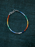Ultra-thin rainbow colored-glaze men’s bracelet with sterling silver clasp, flat lay on dark textured background, minimal identity style