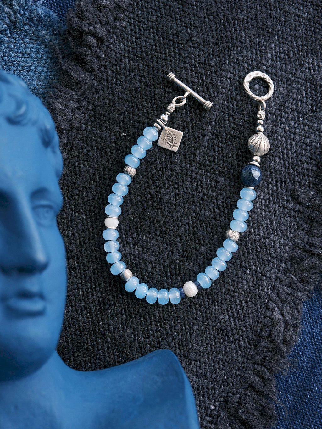 Blue Shoal men’s blue chalcedony bracelet laid on dark woven fabric with sterling silver shell charm and ocean-inspired design