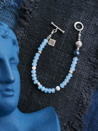 Blue Shoal men’s blue chalcedony bracelet laid on dark woven fabric with sterling silver shell charm and ocean-inspired design
