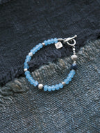 Blue Shoal men’s blue chalcedony bracelet laid on dark woven fabric with sterling silver shell charm and ocean-inspired design