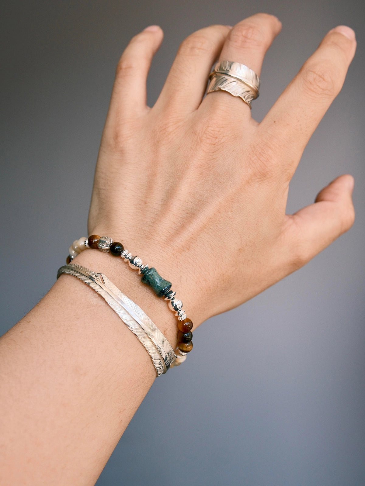 Men’s tiger eye and mother-of-pearl bracelet stacked with a silver feather cuff for a refined quiet-luxury wrist look