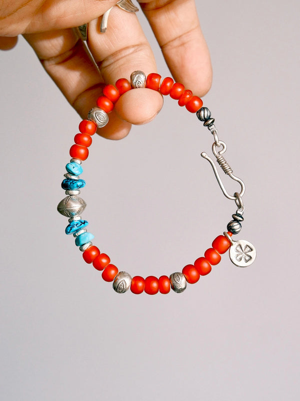 Scarlet Maverick bracelet held between fingers, showing red colored-glaze beads, turquoise accents, a fishhook clasp and a round charm.