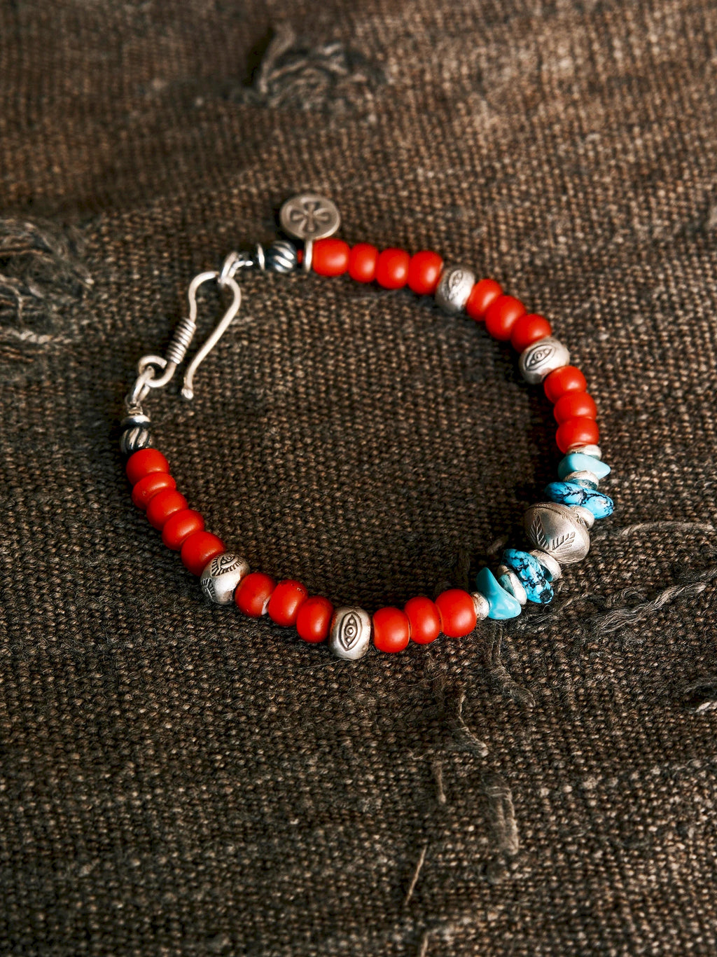 Scarlet Maverick in a circle on textured brown fabric, showing red colored-glaze, turquoise accents, engraved beads and its hook clasp.