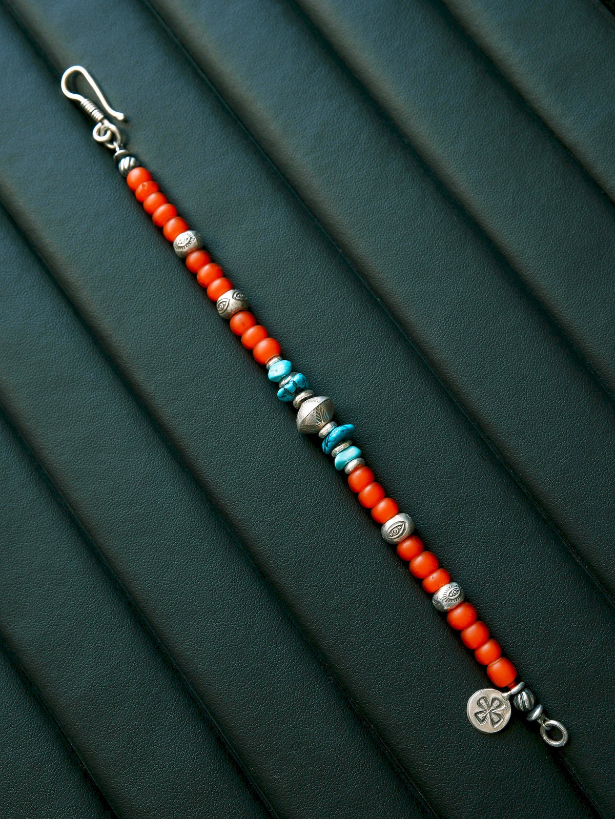 Full Scarlet Maverick bracelet extended diagonally on a dark green textured surface, with turquoise accents, a round charm and an open fishhook clasp.