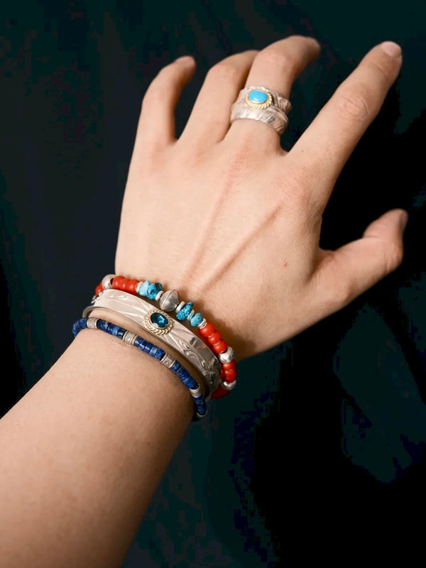 Scarlet Maverick on a wrist against a dark background, styled beside a wide silver-tone cuff, blue beaded bracelet and ring.