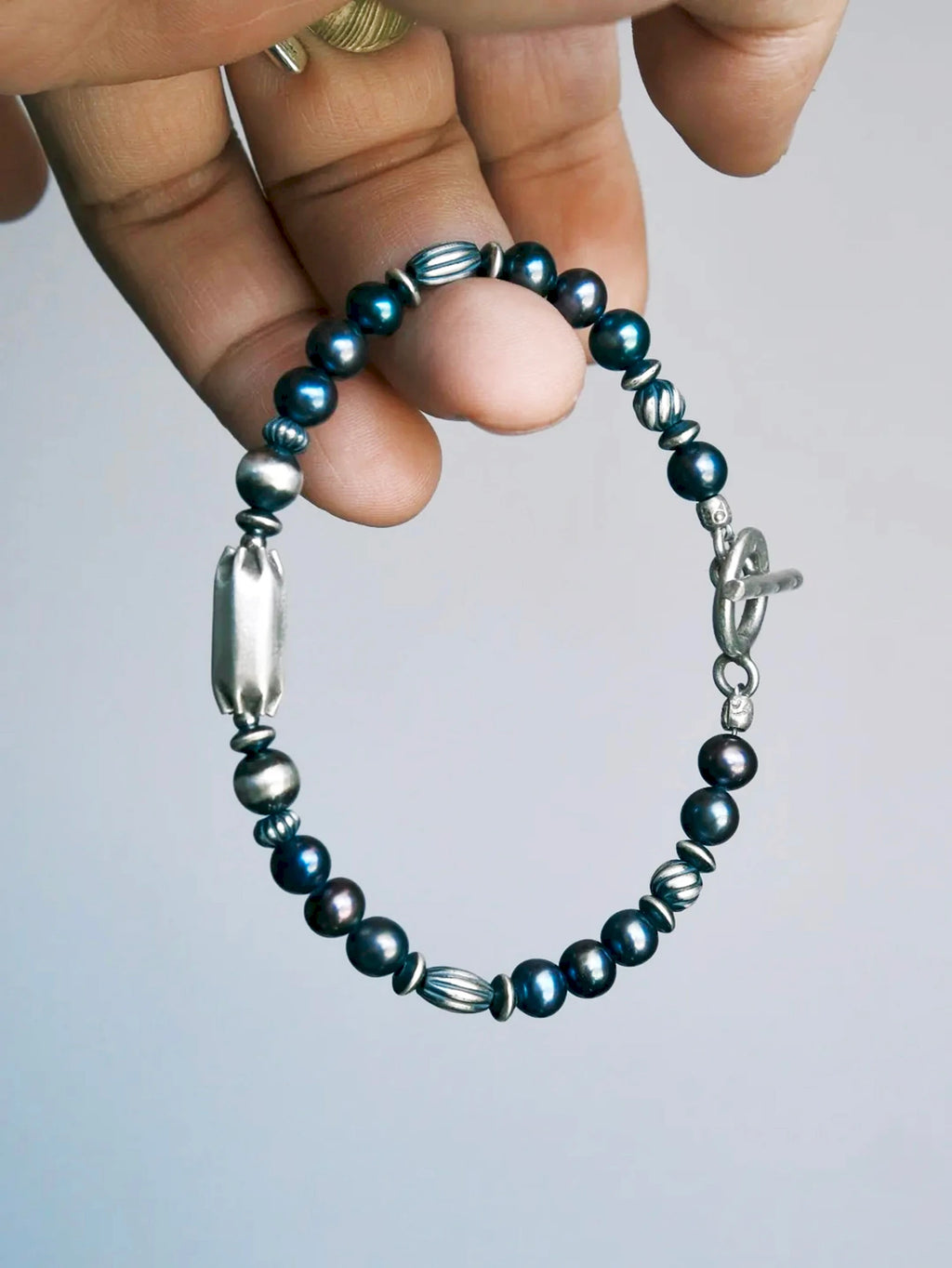 Midnight Envoy held between fingers, showing dark lustrous pearls, ribbed silver beads, an elongated centerpiece and a toggle clasp.