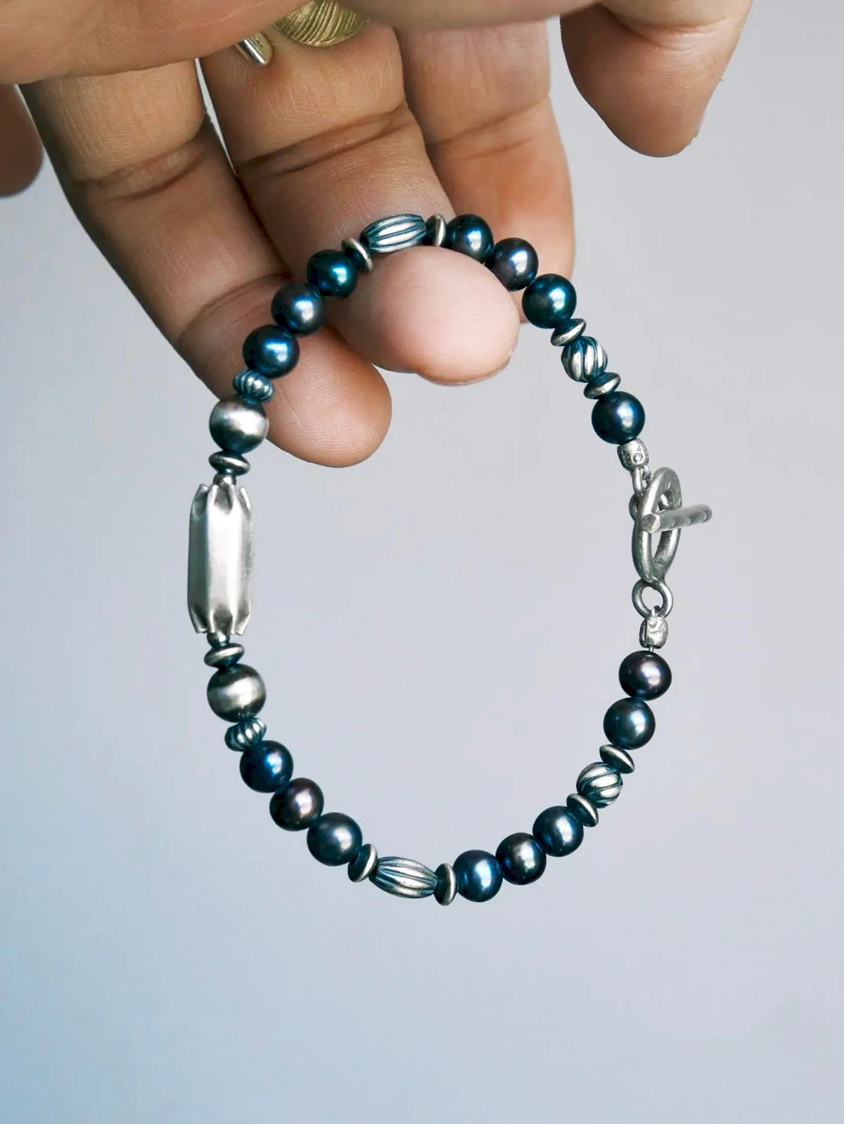 Midnight Envoy held between fingers, showing dark lustrous pearls, ribbed silver beads, an elongated centerpiece and a toggle clasp. Main image