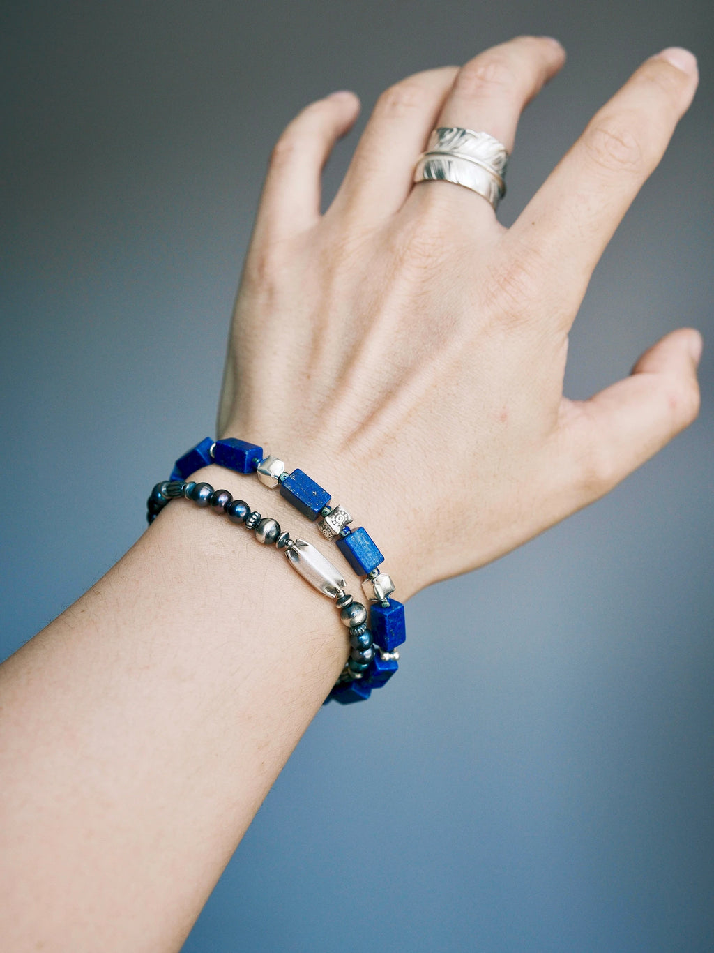 Midnight Envoy pearl bracelet on a wrist with its elongated silver centerpiece facing outward, beside a rectangular blue-bead bracelet.