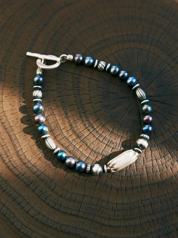Midnight Envoy pearl bracelet in a circle on sunlit wood, with ribbed silver beads, an elongated centerpiece and a closed toggle clasp.