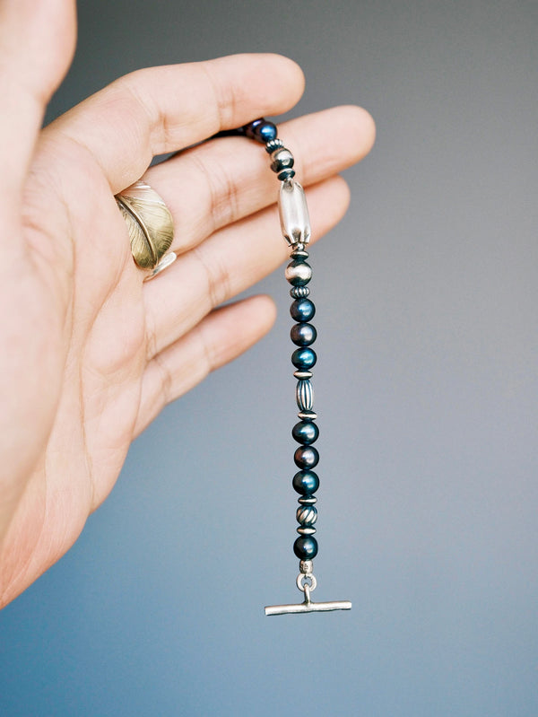 Midnight Envoy held vertically over a palm, showing its elongated silver centerpiece, dark pearls, ribbed beads and toggle bar.
