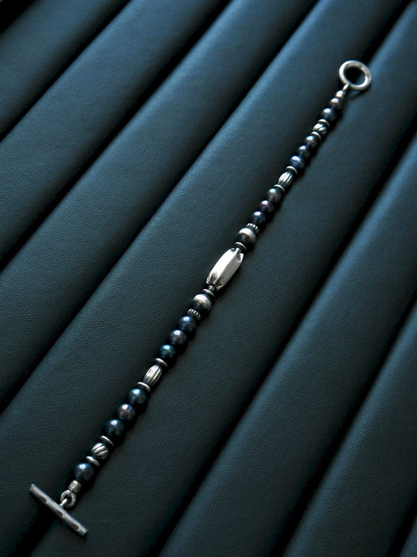 Full Midnight Envoy bracelet extended diagonally on a dark blue textured surface, showing dark pearls, silver details and open toggle ends.