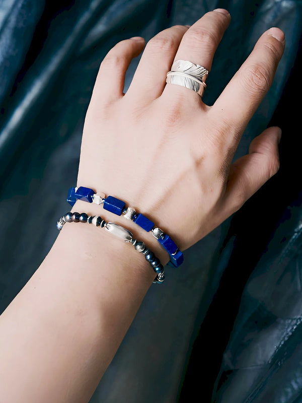 Midnight Envoy pearl bracelet worn beside a chunky rectangular blue-bead bracelet against a dark textured background.