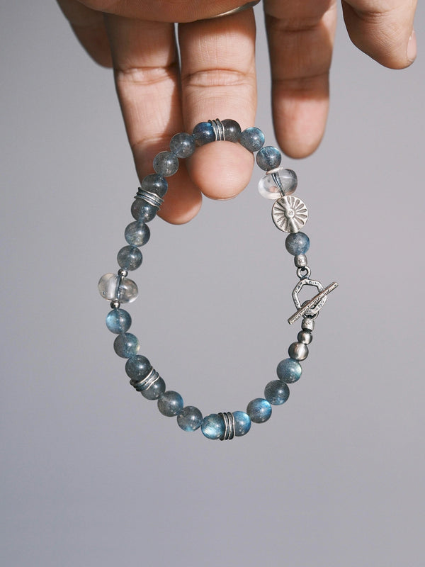 Grey Sovereign held between fingers, with grey labradorite beads, clear quartz accents, a silver sun-motif bead and a geometric toggle clasp.