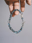 Grey Sovereign held between fingers, with grey labradorite beads, clear quartz accents, a silver sun-motif bead and a geometric toggle clasp.