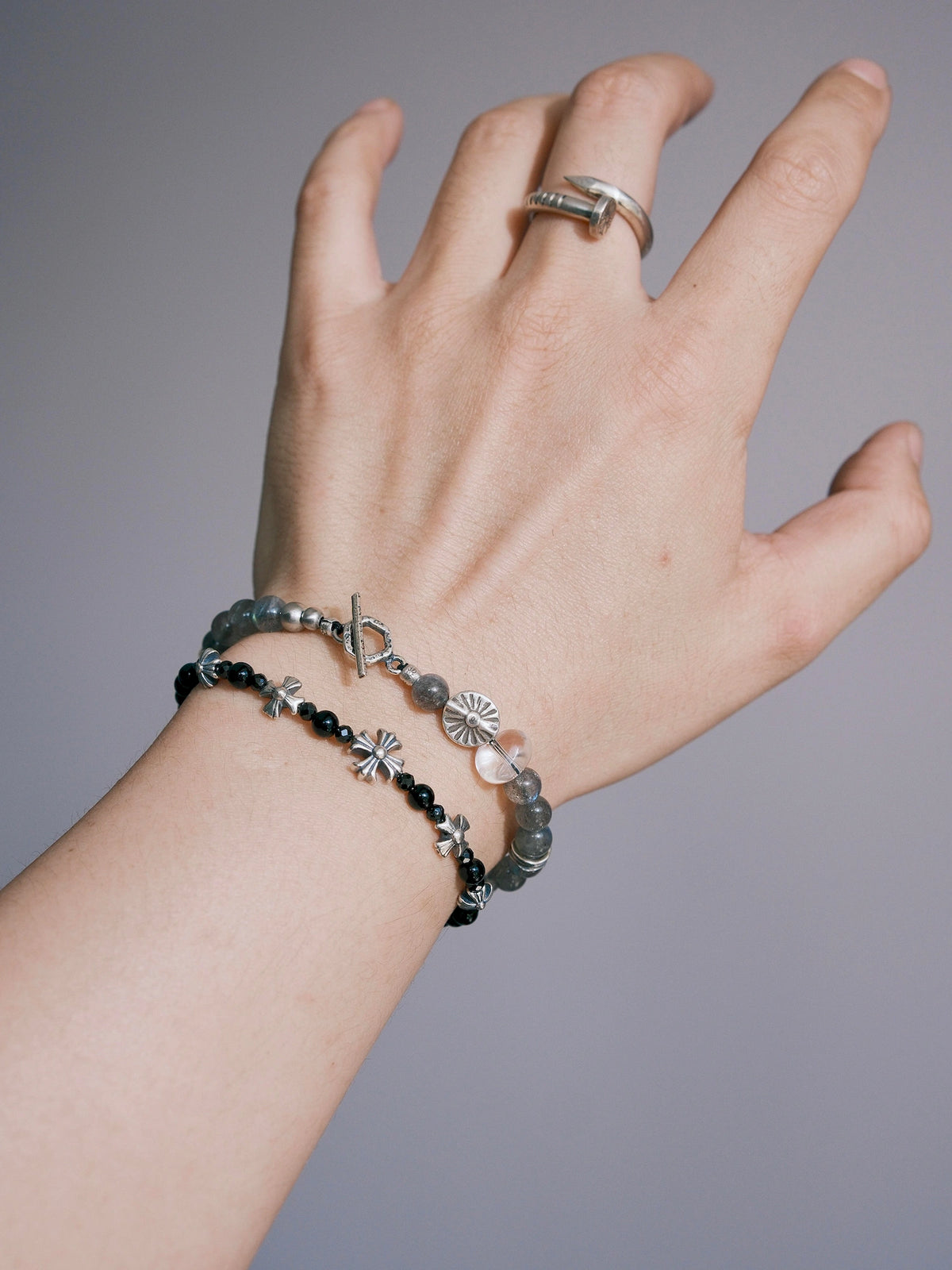 Grey Sovereign on a wrist with its geometric toggle clasp, silver sun-motif bead and clear quartz accent visible beside a black bracelet.