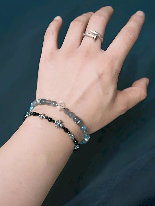 Grey Sovereign wrist close-up showing blue flashes in grey labradorite, a clear quartz accent and silver spacers beside a black-bead bracelet.