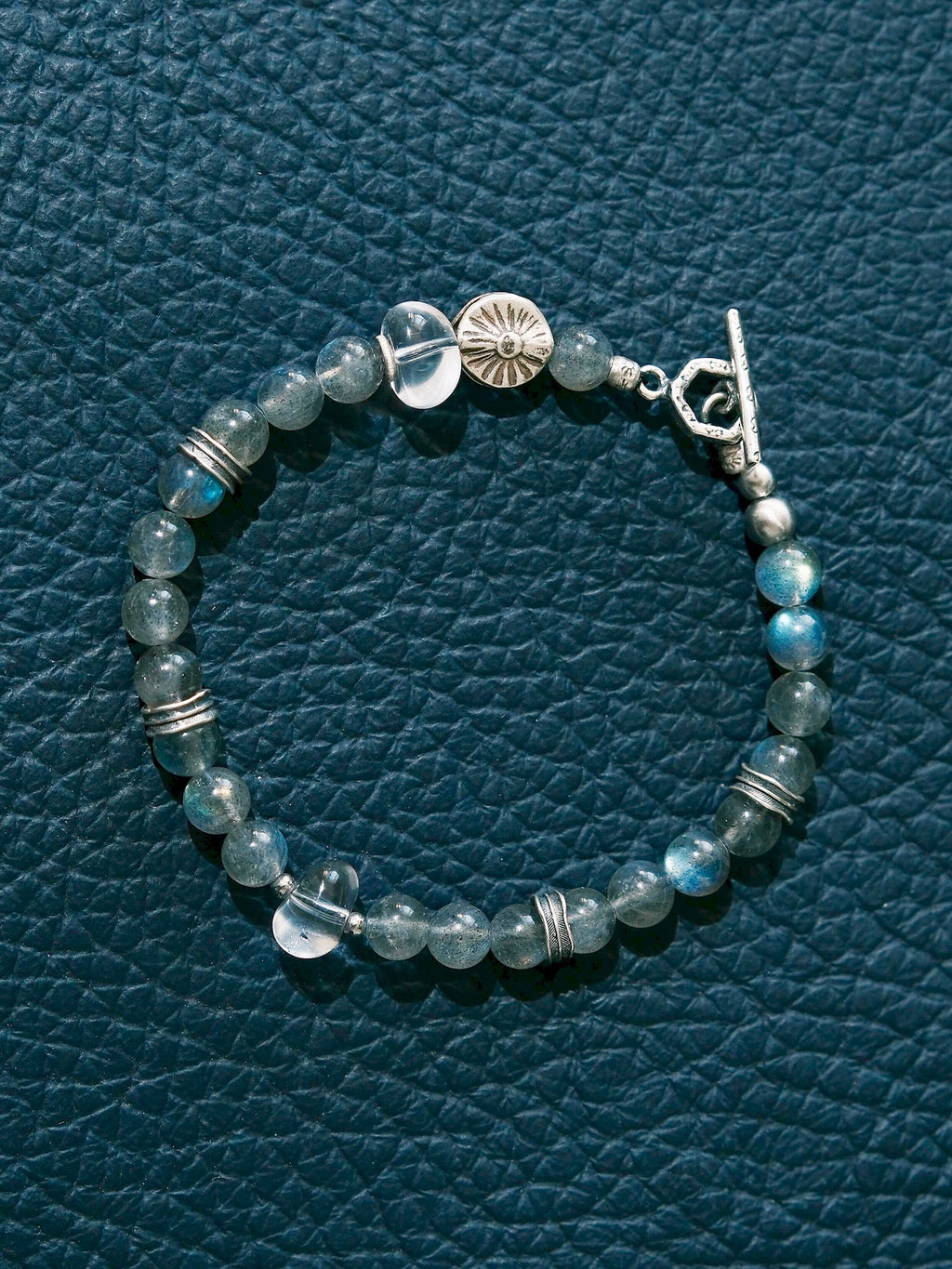 Grey Sovereign laid in a circle on a dark blue textured surface, with labradorite, clear quartz, a silver sun-motif bead and toggle clasp.
