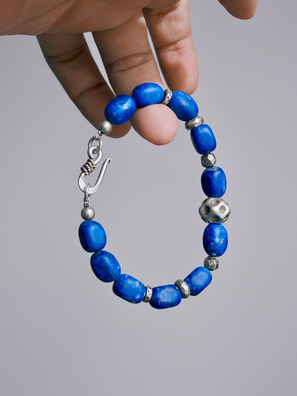 Lapis lazuli bracelet held against a gray background, showing blue barrel-shaped beads, silver spacers and a hook clasp. Main image