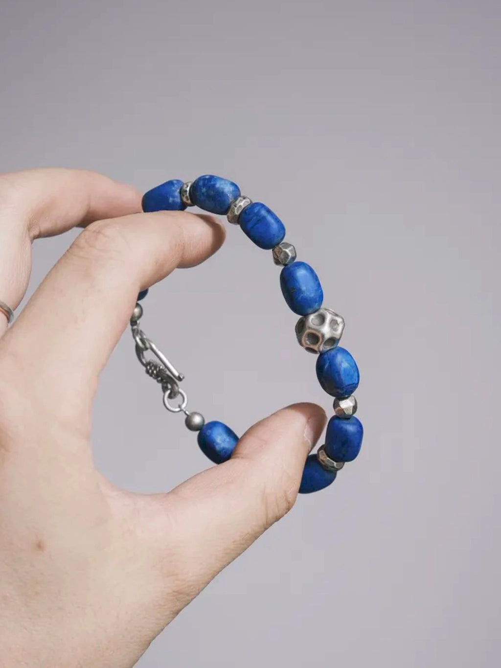 Hand holding a closed lapis lazuli bracelet, with the cratered silver bead and hook clasp visible.