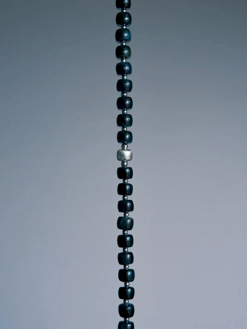 Cropped vertical detail of Blue Strategist dark blue beads and a larger textured silver-tone bead on gray.
