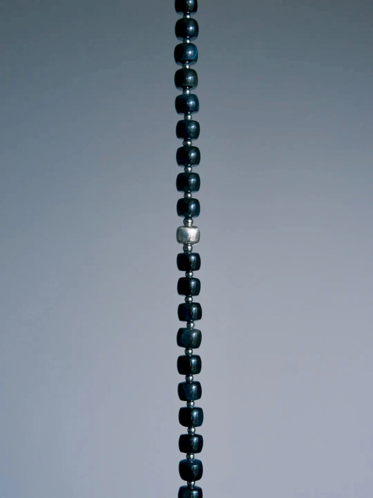 Cropped vertical detail of Blue Strategist dark blue beads and a larger textured silver-tone bead on gray.