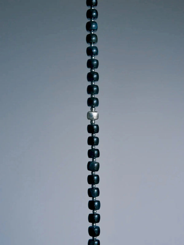 Cropped vertical detail of Blue Strategist dark blue beads and a larger textured silver-tone bead on gray.