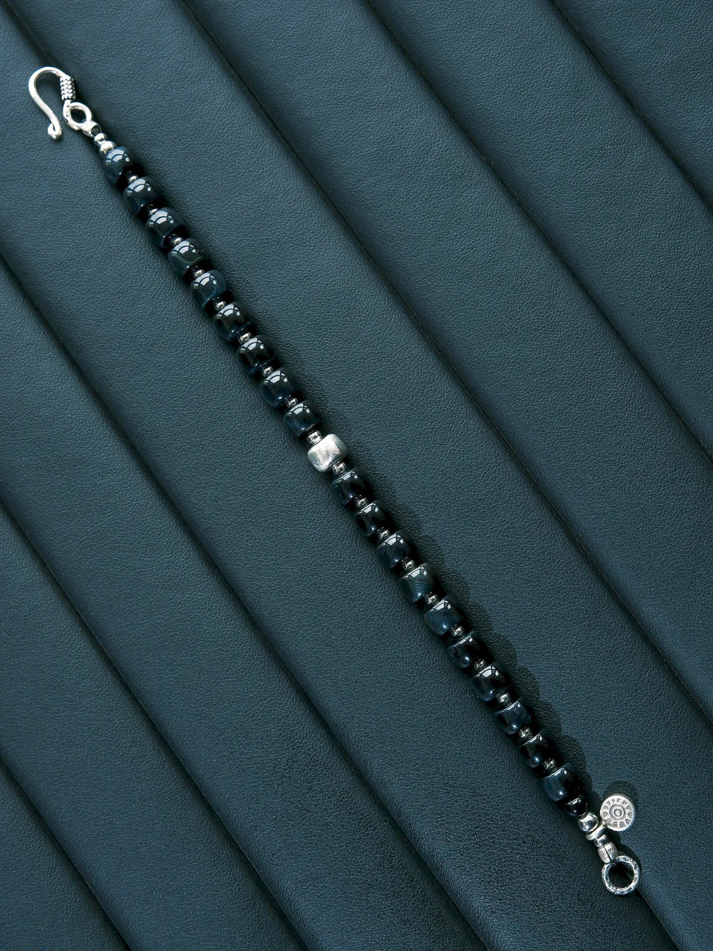 Blue Strategist bracelet laid open diagonally on a dark padded surface, with a hook clasp and round end ring.