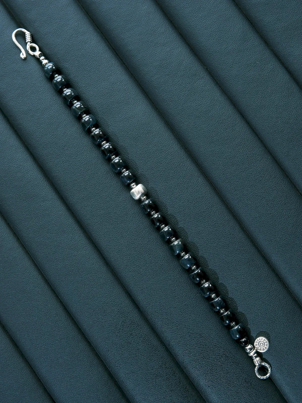Blue Strategist bracelet laid open diagonally on a dark padded surface, with a hook clasp and round end ring.
