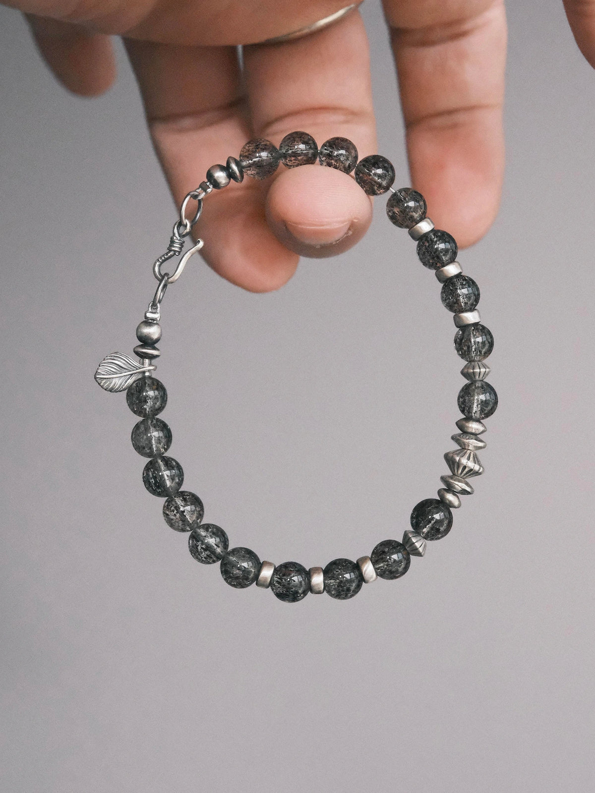 Shadow Sentinel bracelet held in hand, showing translucent dark beads, a fishhook clasp and a leaf-shaped detail. Main image