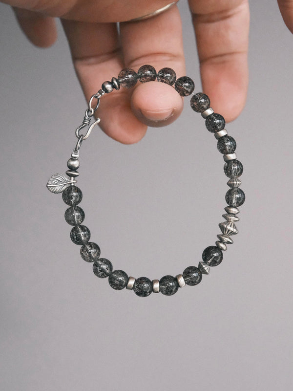 Shadow Sentinel bracelet held in hand, showing translucent dark beads, a fishhook clasp and a leaf-shaped detail.
