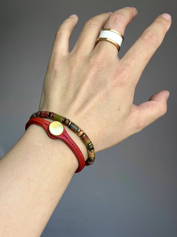 Gold-tone Earth Nomad bracelet worn beside a red leather bracelet, with a white-and-gold ring.