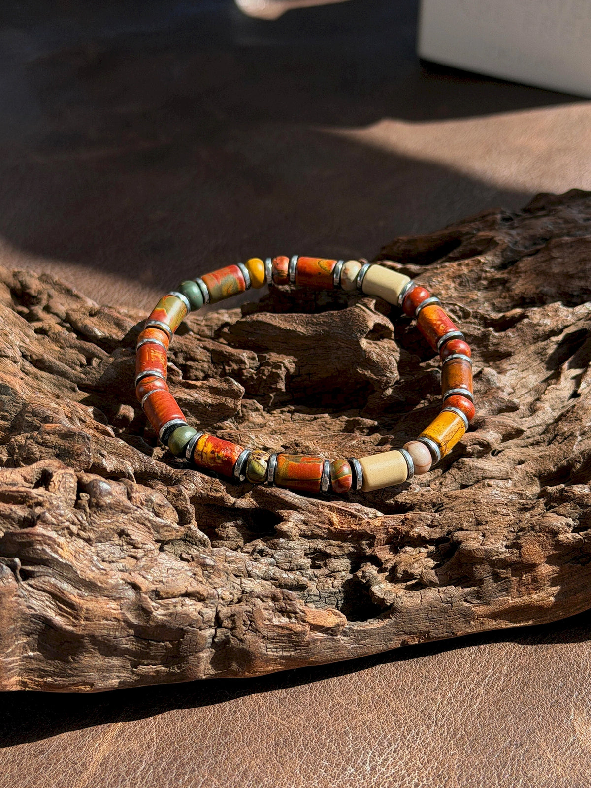 Silver-tone Earth Nomad bracelet on textured bark in sunlight, showing red, green and beige stone beads.