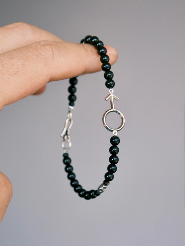 Silver Ego black onyx bracelet held against a gray background, showing the silver male symbol and fishhook clasp.